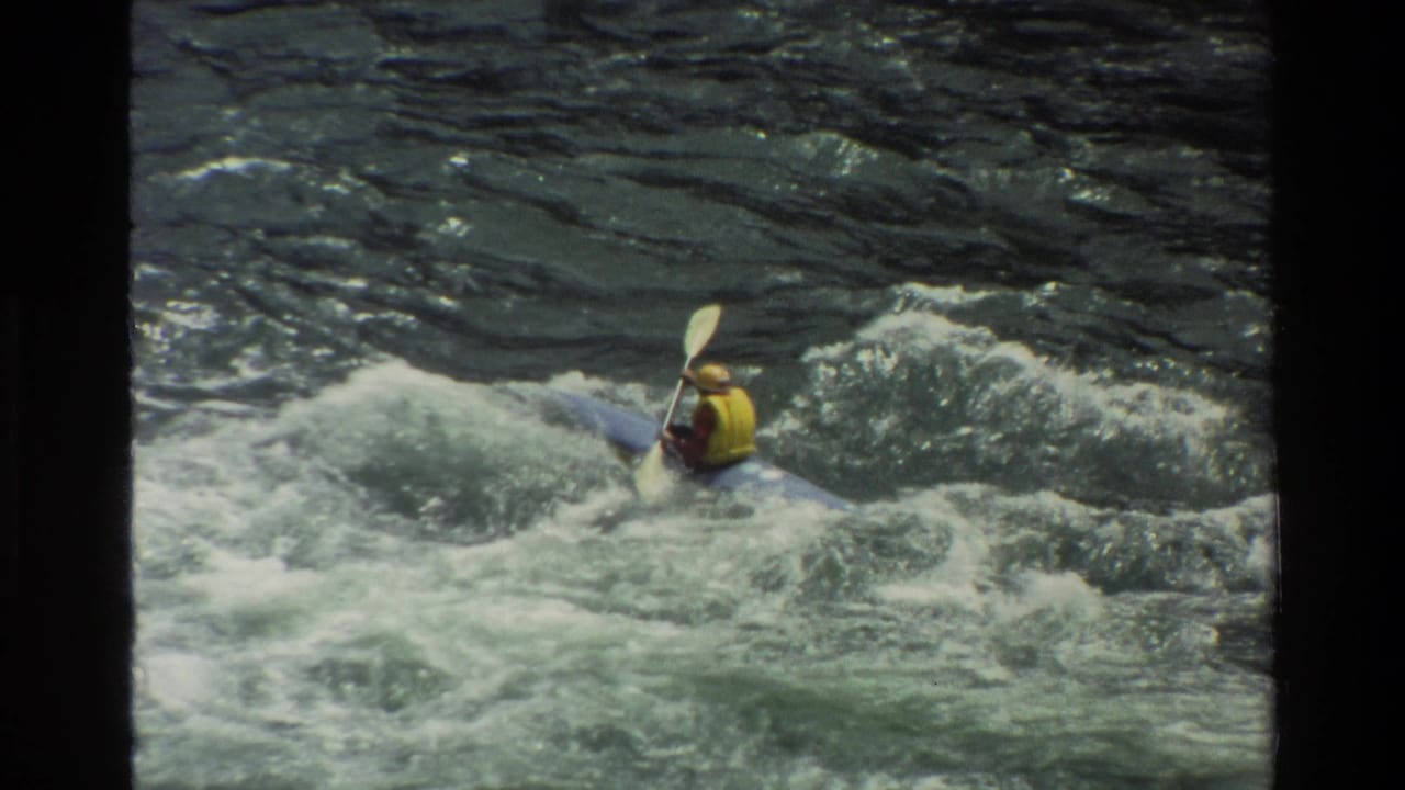 Kayaker paddles across a cresting wave in rough river water.