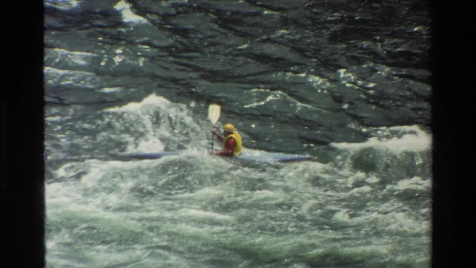 A distant kayaker in safety gear is partly hidden by spray and rough water in this 1980 Wyoming river scene.
