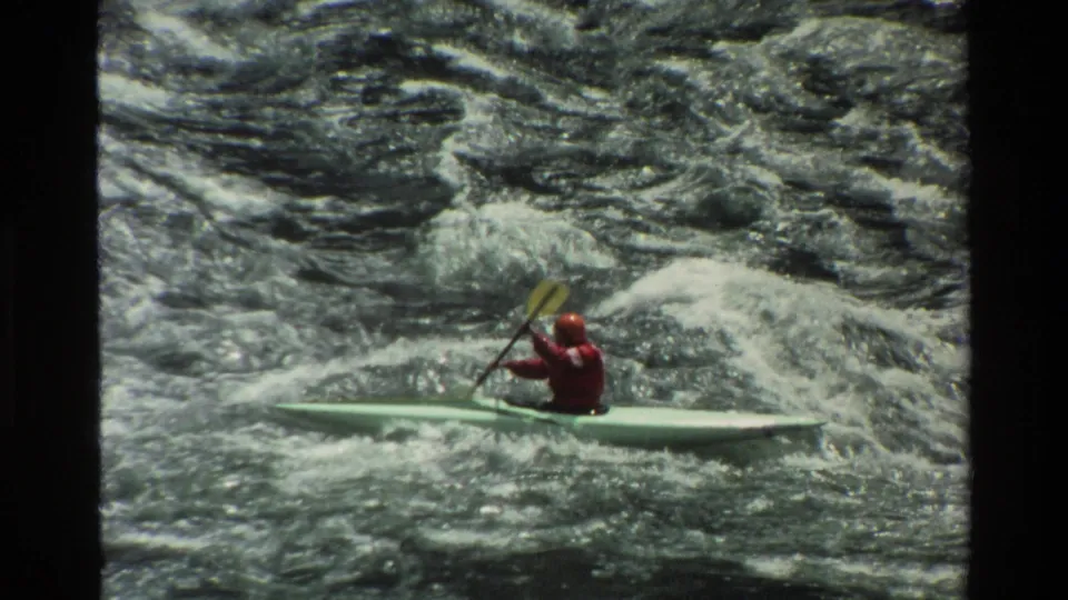A second or differently framed paddler in red gear moves across foaming river water in a light green kayak.