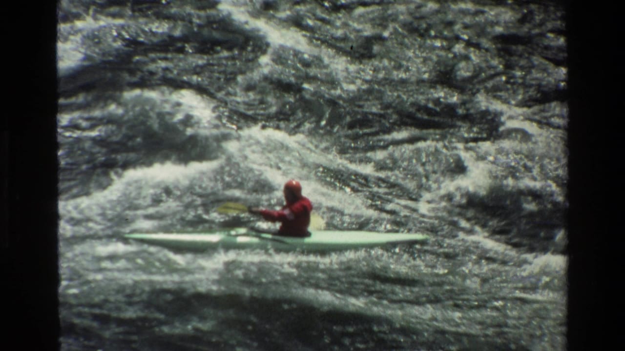 1980 Grand Teton home-movie frame showing a kayaker in red gear moving across churning whitewater in a pale kayak.