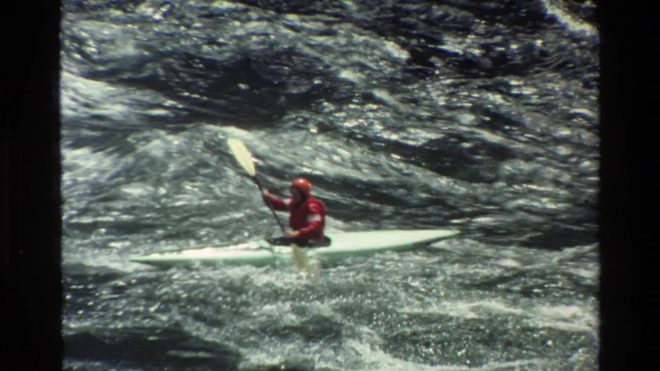 1980 Grand Teton home-movie frame with a red-clad kayaker steering a pale kayak through foamy whitewater.