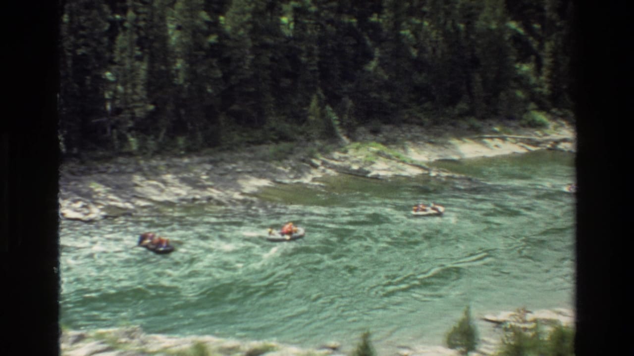 1980 Grand Teton home-movie frame showing distant rafters or tubers on a green-blue river below forested banks.