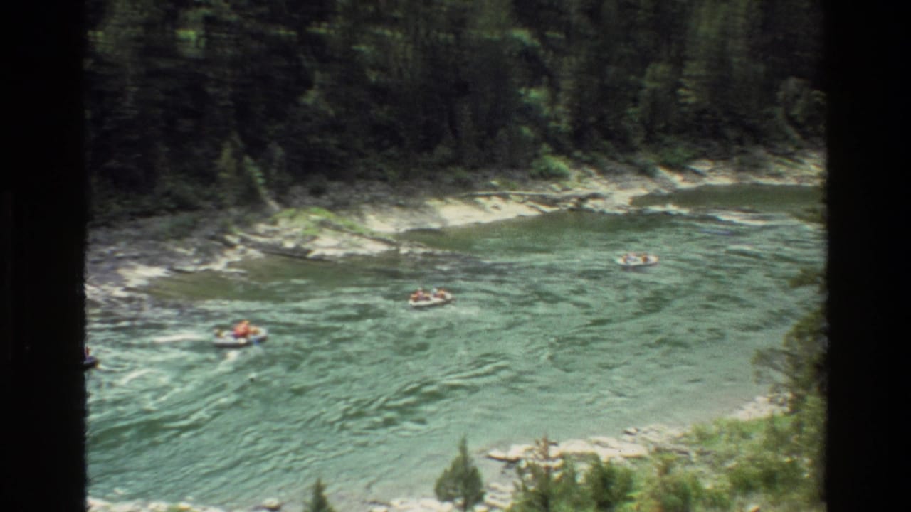 Small rafts or tubes drift across a wide river with dense trees and rocky shoreline behind them.