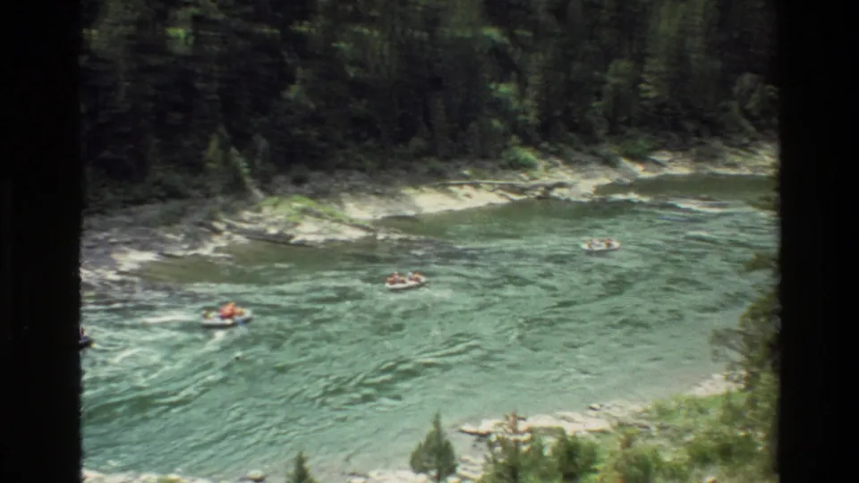 1980 Grand Teton footage frame with distant people floating on a river beneath a dark evergreen forest edge.