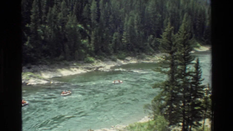 High vantage 1980 Grand Teton frame showing a forested river bend with distant recreation craft in the water.