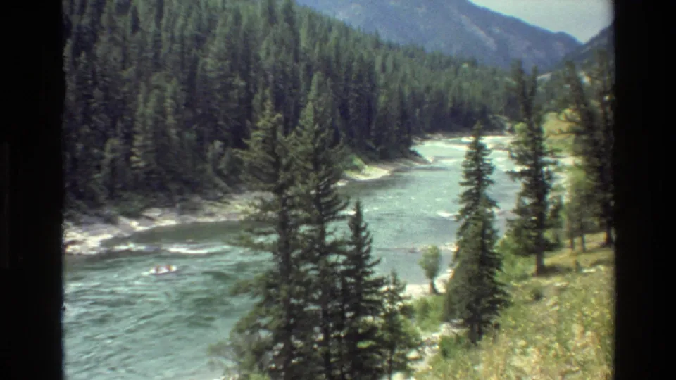 Vintage 8mm view of a Grand Teton, Wyoming river corridor, with foreground evergreens and a distant raft carrying people.