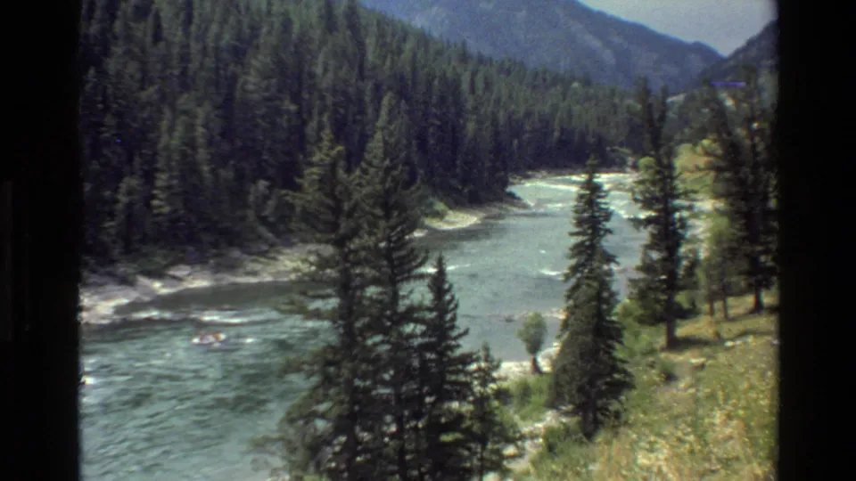 1980 home-movie frame from Grand Teton, Wyoming showing a broad mountain river, pine forest, and a small distant raft.
