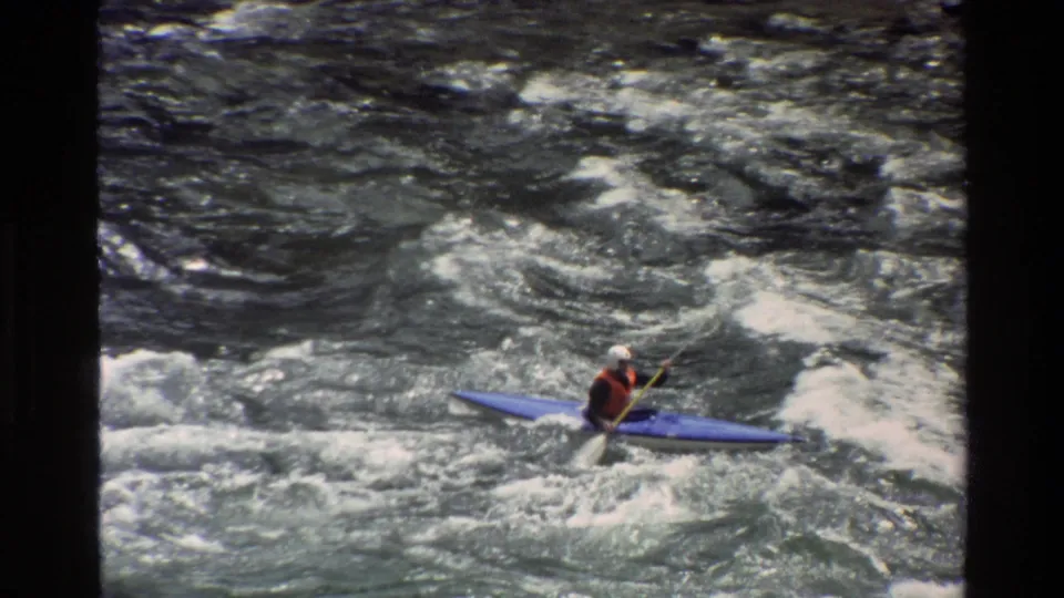 1980 8mm frame from Grand Teton, Wyoming showing a helmeted kayaker moving across foamy whitewater in a blue kayak.