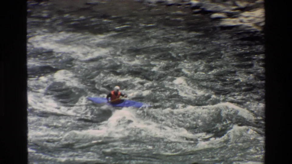 In 1980 Grand Teton, Wyoming footage, a kayaker in a blue kayak paddles across churning whitewater seen from above.