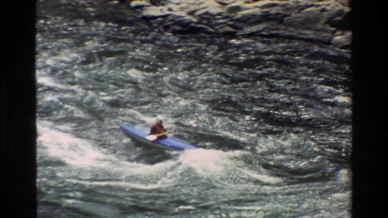 Kayaker in a blue kayak riding through rough water near a rocky river edge.