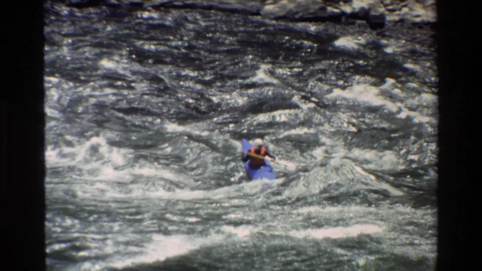 A high-angle 8mm frame shows the kayaker lower in the shot amid swirling whitewater and dark river currents.