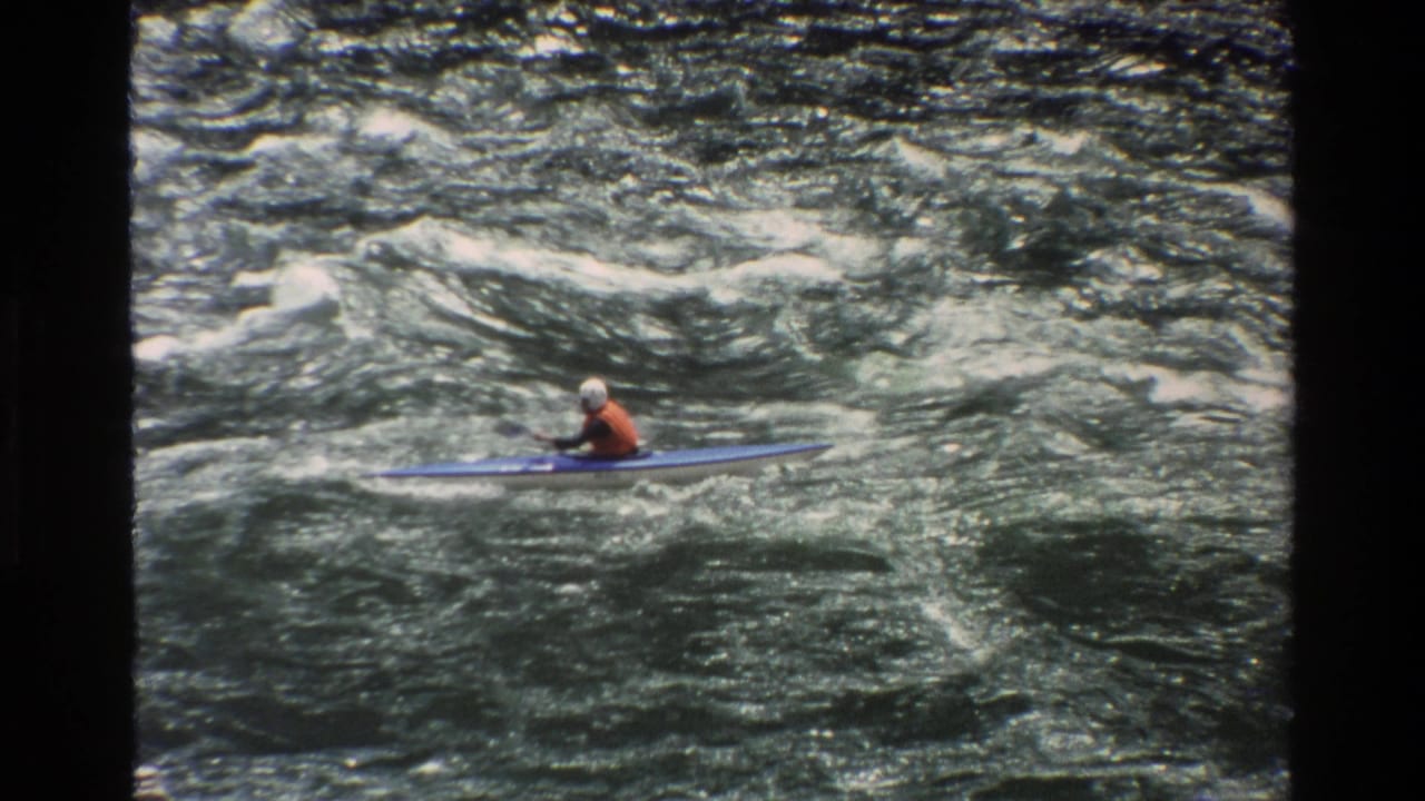 A distant kayaker in helmet and life vest paddles across rough whitewater in Grand Teton, Wyoming footage from 1980.