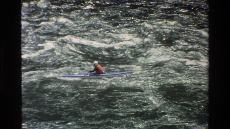 A distant kayaker in helmet and life vest paddles across rough whitewater in Grand Teton, Wyoming footage from 1980.