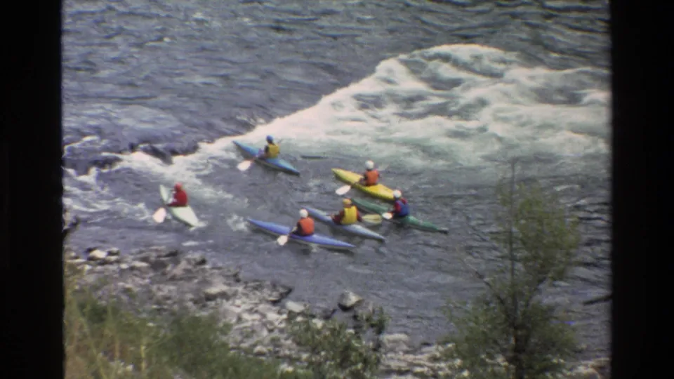 Several kayakers in colorful boats gather near a rocky bank beside whitewater in Grand Teton, Wyoming footage from 1980.