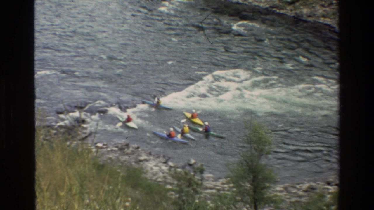 Kayakers move along the river edge near a foamy rapid.