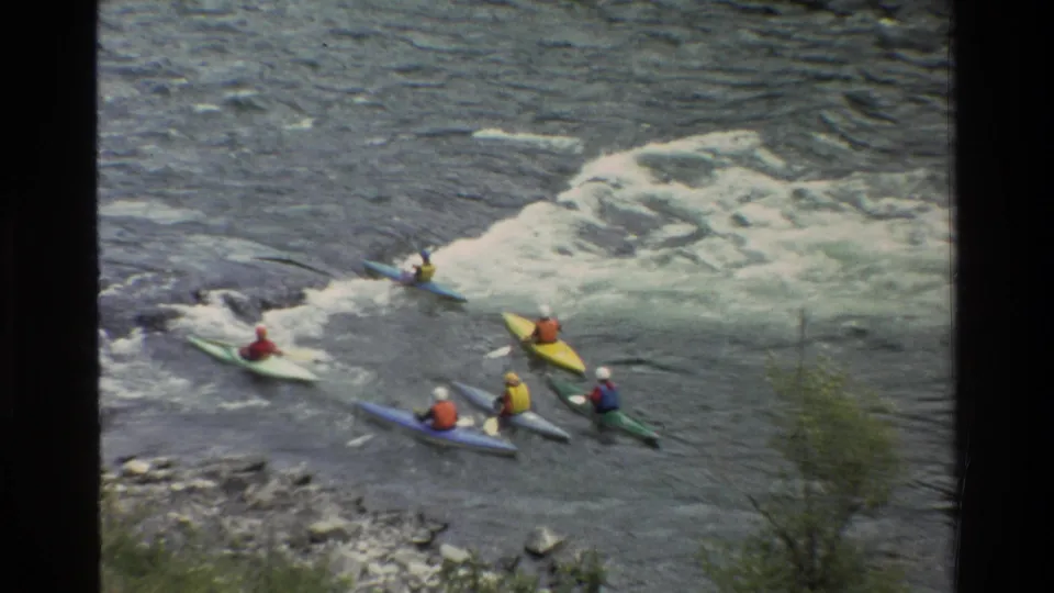 Six helmeted kayakers in bright boats paddle downstream beside whitewater and a rocky riverbank.