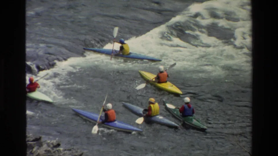Kayakers in helmets and life vests approach a foamy rapid, filmed from above in 1980 Wyoming home-movie footage.