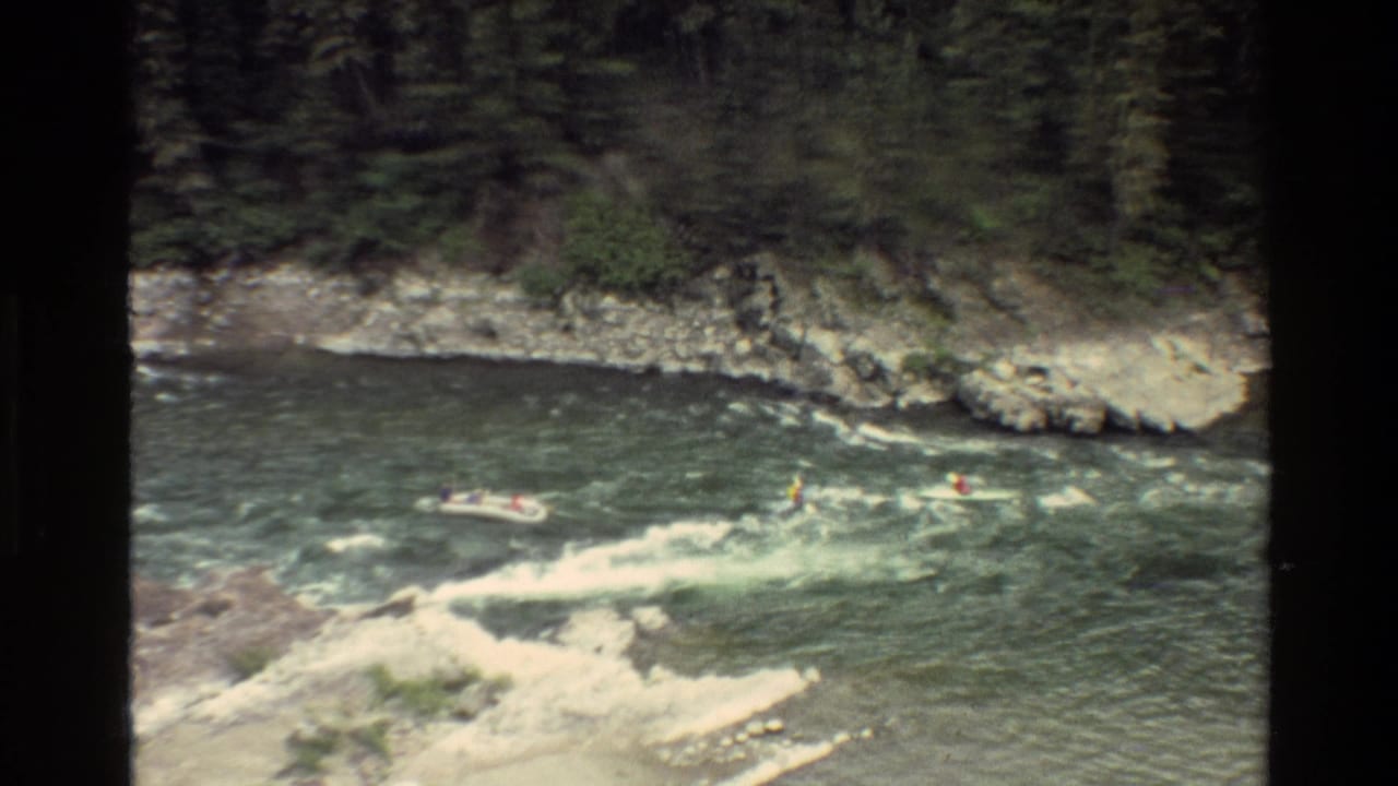 1980 Grand Teton home-movie frame showing a raft and kayakers in green white water below a rocky evergreen bank.