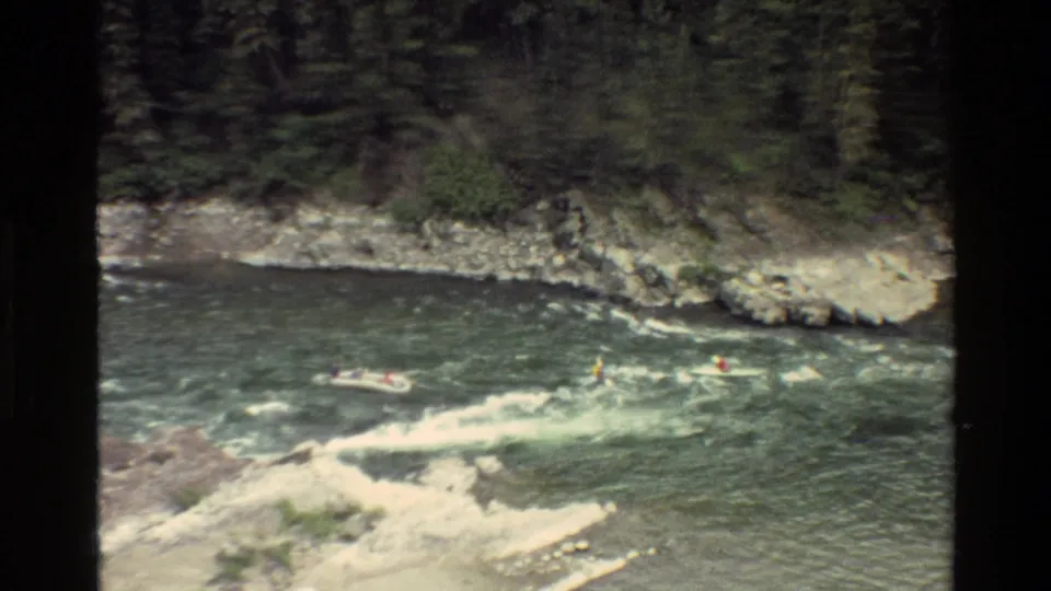 1980 Grand Teton home-movie frame showing a raft and kayakers in green white water below a rocky evergreen bank.