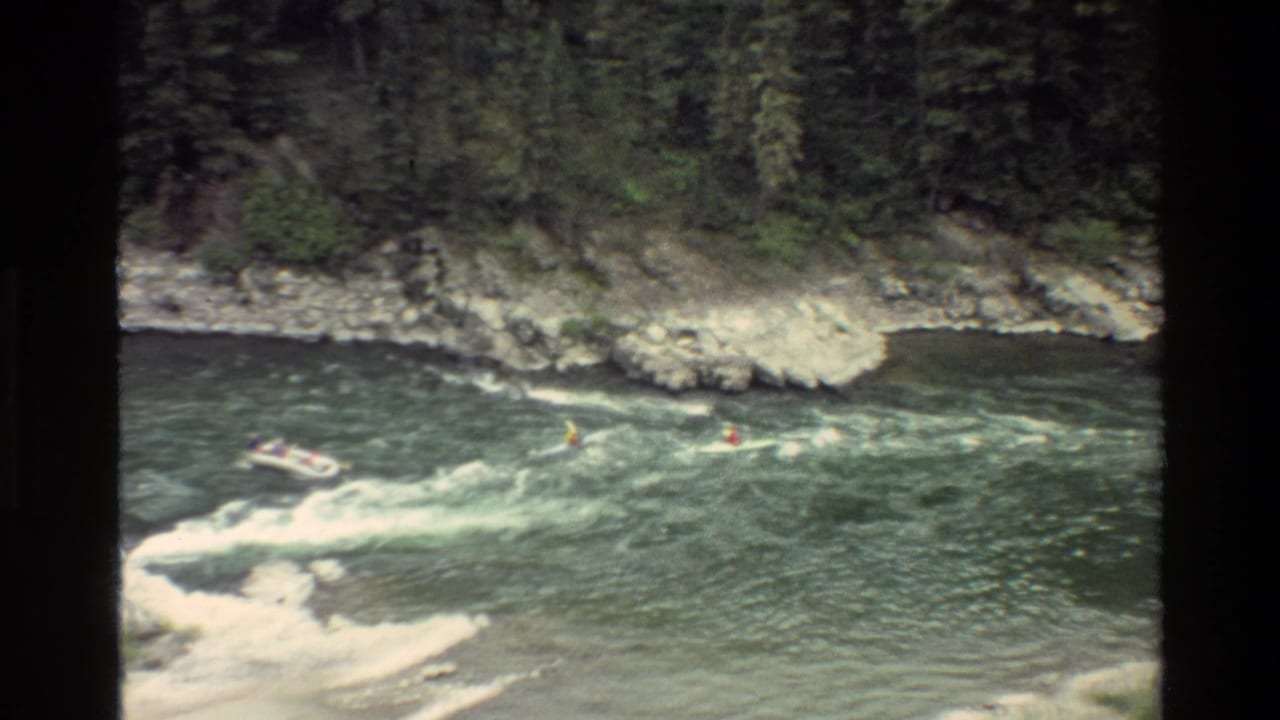 Raft and kayakers continue downstream through rapids near a wooded rocky bank.