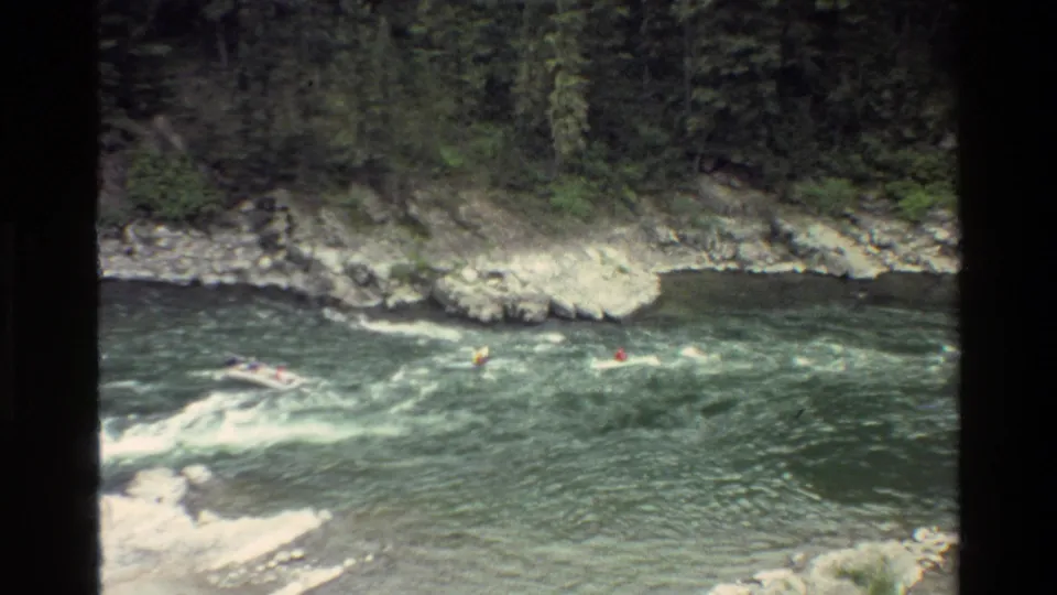 Distant 1980 Grand Teton home-movie view of kayakers and a raft moving through white water along a rocky bank.