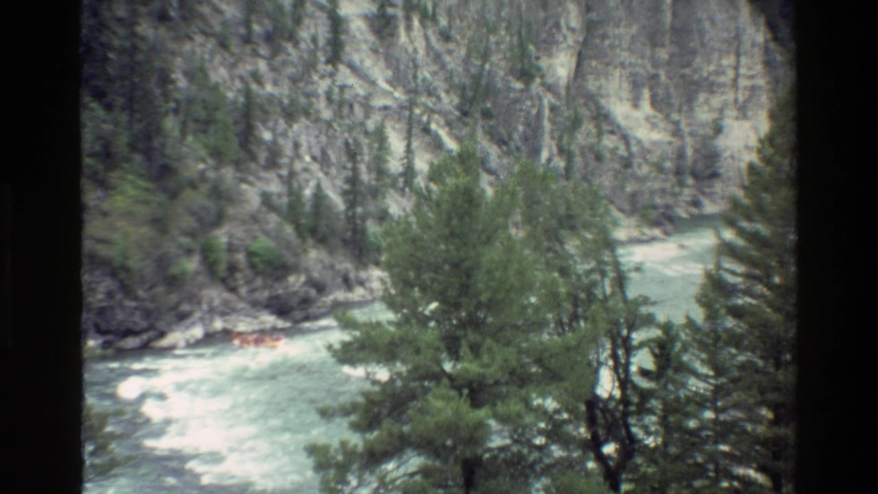 1980 8mm view of a small orange raft in turbulent water below rocky Grand Teton canyon walls and pines.