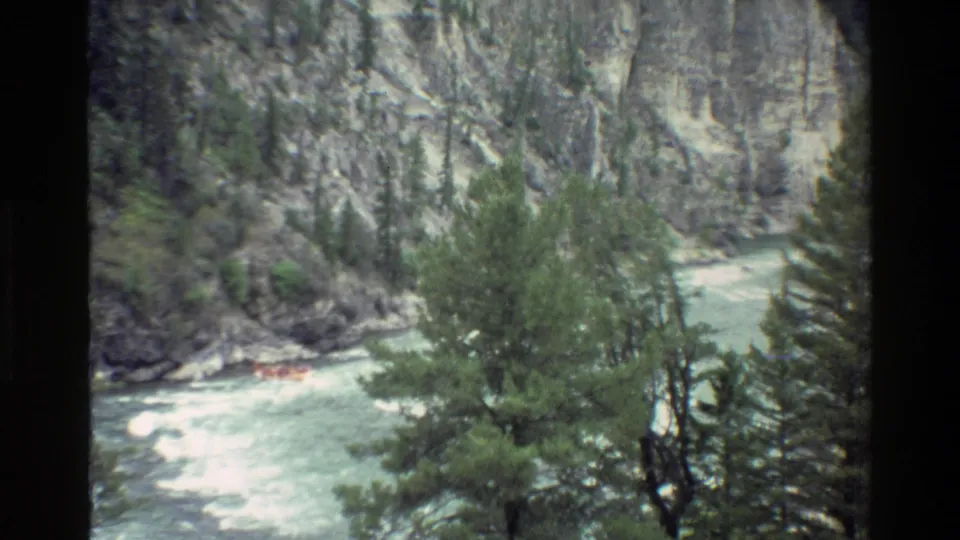 1980 8mm view of a small orange raft in turbulent water below rocky Grand Teton canyon walls and pines.