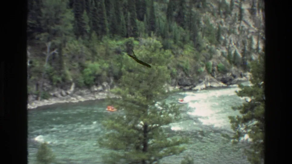 Orange raft passes through a canyon river scene, partly screened by trees in grainy 1980 Grand Teton 8mm footage.