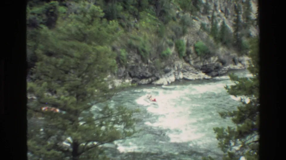 A small raft appears near foaming rapids in a forested canyon river, viewed from above in 1980 Wyoming 8mm footage.