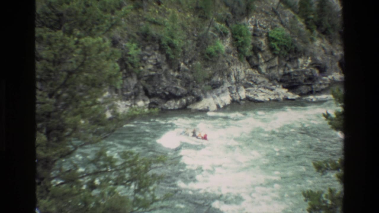 1980 8mm home-movie frame from Grand Teton, Wyoming, showing a distant raft entering churning rapids in a rocky, wooded river corridor.