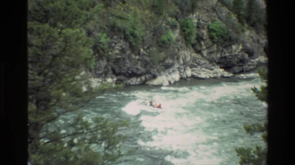 1980 8mm home-movie frame from Grand Teton, Wyoming, showing a distant raft entering churning rapids in a rocky, wooded river corridor.