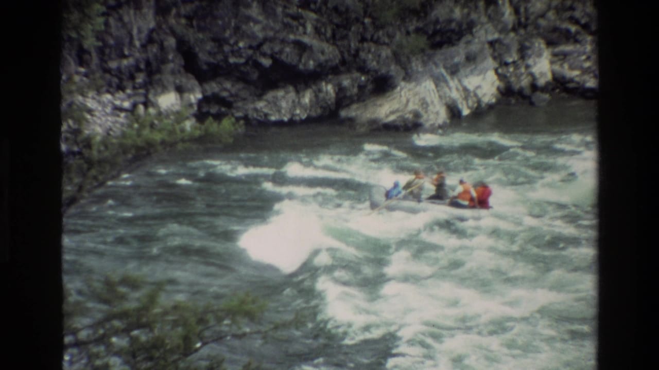 Raft with several people in life jackets moves through foamy rapids near rock walls.