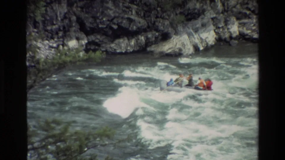 1980 Grand Teton rafting footage shows several paddlers in bright life jackets riding a raft through foamy whitewater beside a rocky bank.