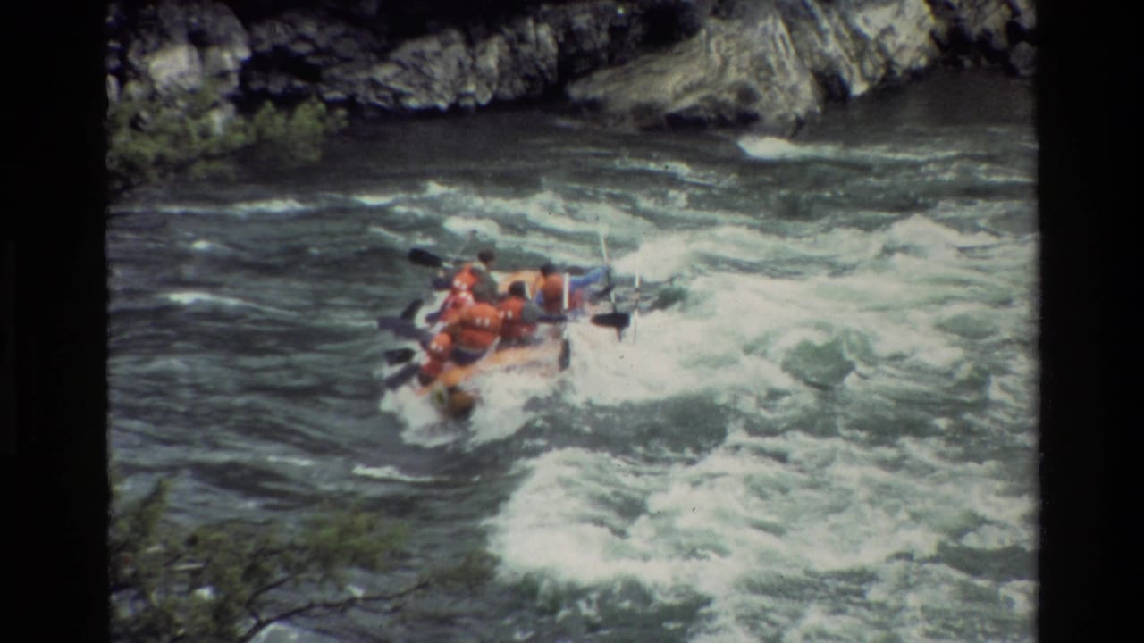 Raft rides over foamy rapids with paddles raised and moving.