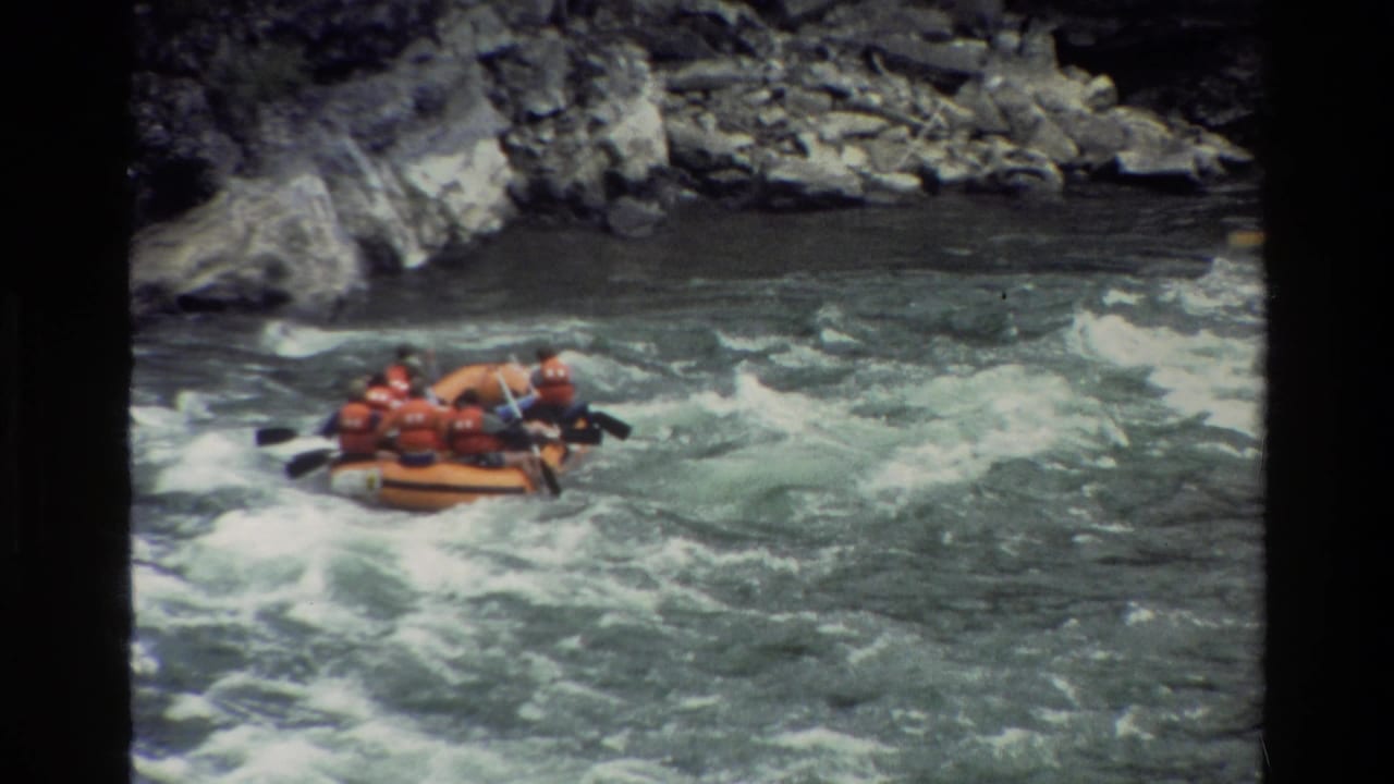 In 1980 Grand Teton rafting footage, an orange inflatable raft carries people in red life vests through turbulent water near rocks.