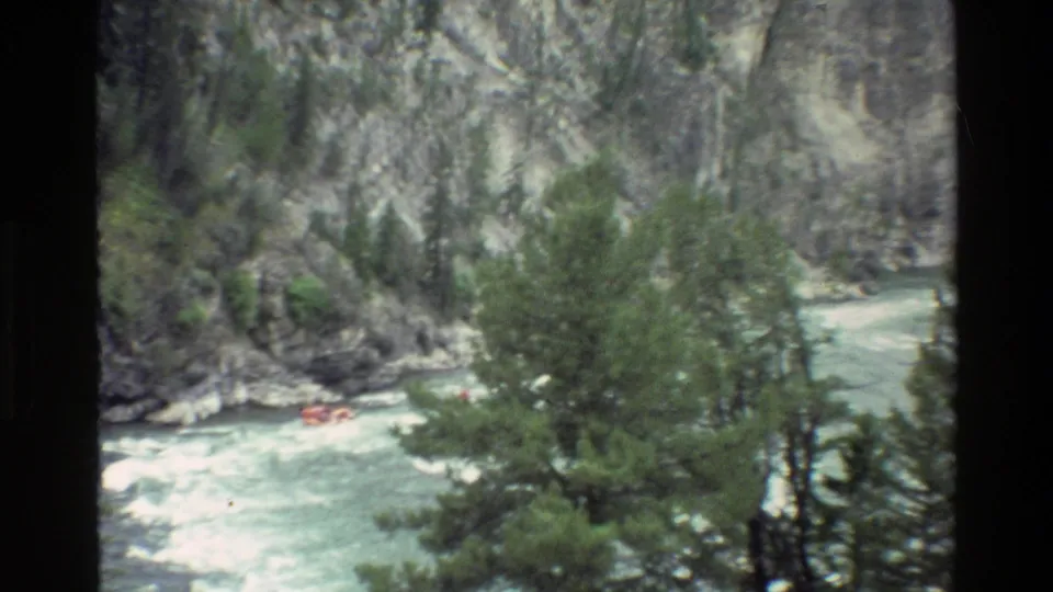 A distant orange raft is visible through trees as it rides the rapids below steep rocky slopes in the Wyoming canyon setting.