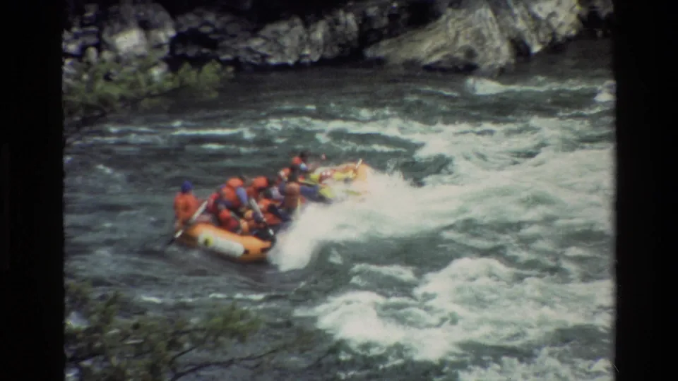 The raft crests a foaming rapid, with several distant rafters bracing and paddling through the turbulent river.