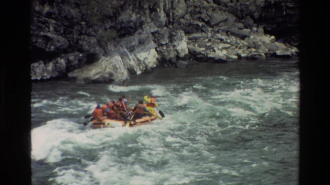 Raft moves across turbulent water with paddlers clustered inside and rocks behind.