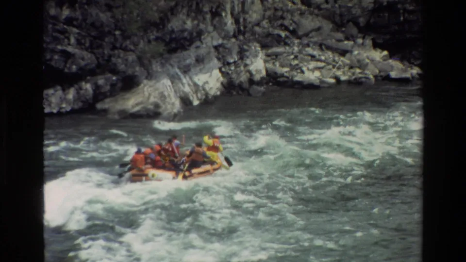 Orange rafting boat continues through Grand Teton river rapids, with distant paddlers and rocky canyon scenery visible.