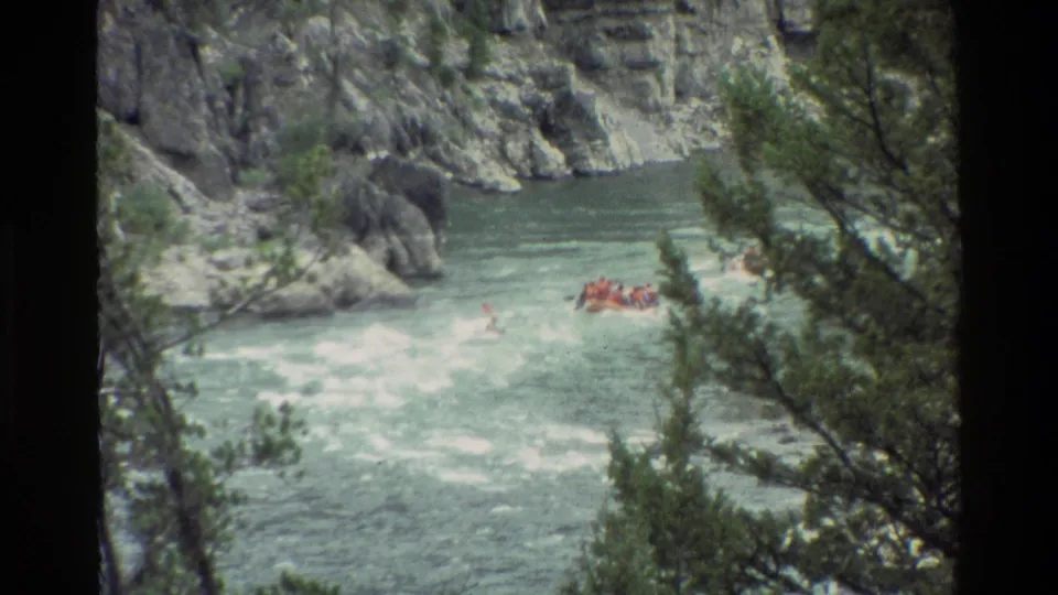 Wide 8mm view of a small orange raft on a turbulent Wyoming river, partly framed by evergreen branches.