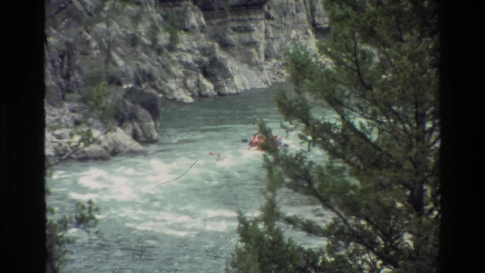 1980 8mm Grand Teton rafting scene with a red raft in rapids, rocky canyon walls, and evergreen trees partly obscuring the view.