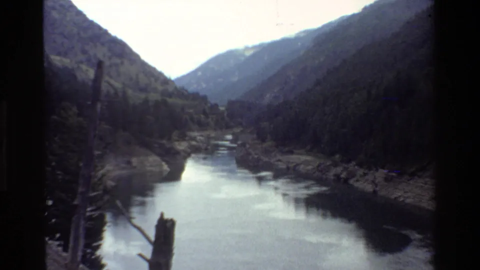 A scenic Grand Teton river valley appears in soft 8mm color, with dark wooded slopes, reflective water, and mountain ridges in the distance.