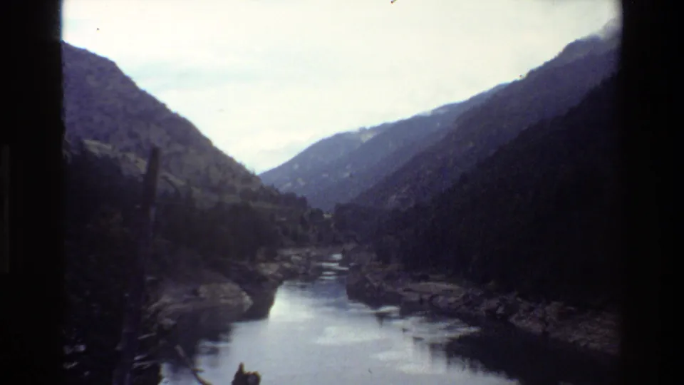 1980 Grand Teton home-movie frame showing a river valley receding between wooded mountain slopes with faded color and dark edge falloff.