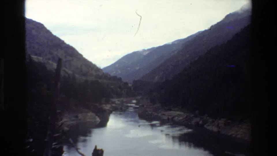 1980 8mm Grand Teton scenic frame with calm water, dark forested slopes, distant ridges, and small visible film marks.