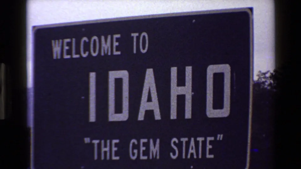 Circa-1980 8mm roadside view of a Welcome to Idaho sign reading The Gem State, with trees at the edge of frame.