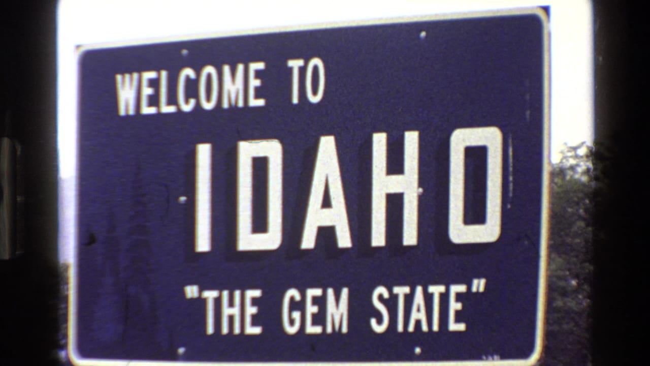 1980 8mm frame showing a blue Welcome to Idaho The Gem State sign with white lettering beside roadside trees.