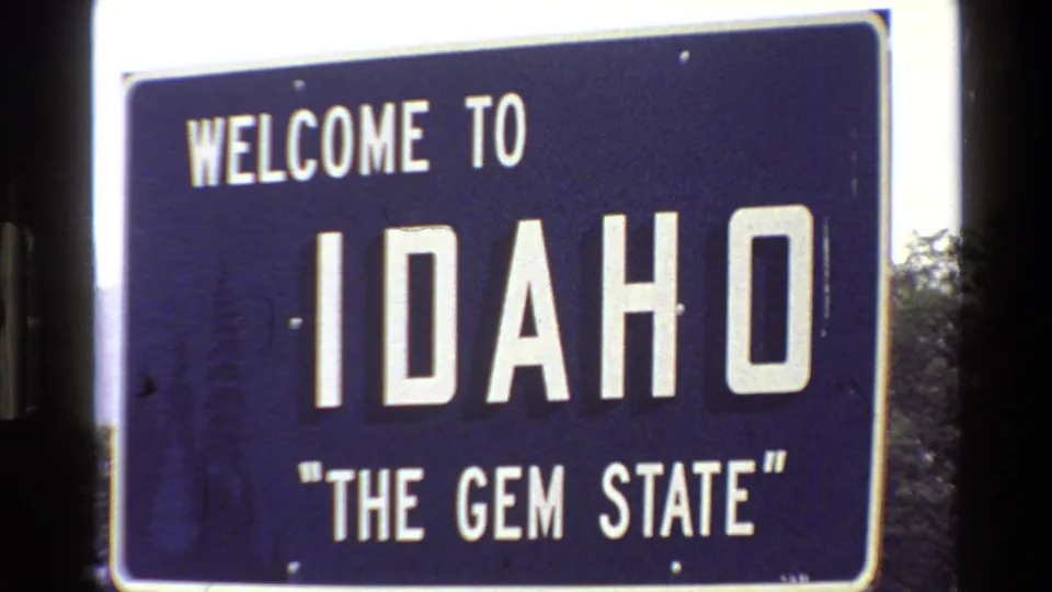 1980 8mm frame showing a blue Welcome to Idaho The Gem State sign with white lettering beside roadside trees.