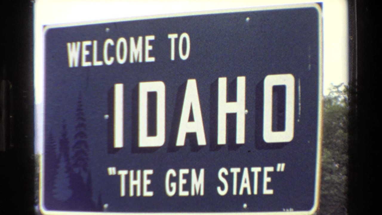Close 1980 8mm view of the Idaho welcome sign, with white block letters and dark trees behind the sign edge.