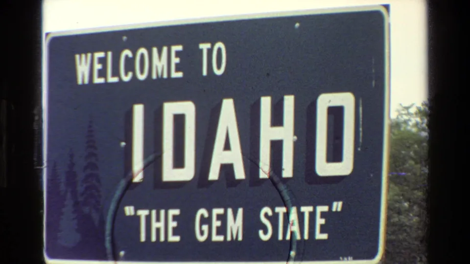 1980 8mm frame with the Idaho welcome sign partly cropped, showing scratches, soft focus, and roadside foliage.