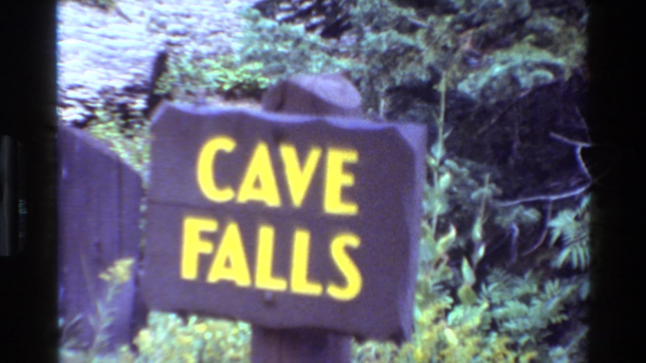 1980 8mm frame of a brown Cave Falls sign with yellow lettering near evergreen forest in the Wyoming travel clip.